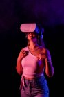 Невпізнавана жінка в гарнітурі VR, що взаємодіє з віртуальною реальністю, стоячи в темній студії з паровим і синім і рожевим неоновим освітленням — стокове фото