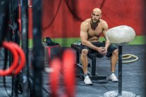 Vista lateral del macho musculoso agotado mirando hacia otro lado sentado sobre pesas y descansando durante el entrenamiento funcional en el gimnasio - foto de stock
