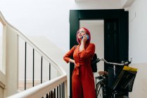 Junge stylische Frau im roten Anzug mit Rucksack spricht auf Smartphone, während sie mit Fahrrad auf der Treppe steht — Stockfoto