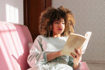 Konzentrierte Afroamerikanerin sitzt zu Hause auf der Couch und liest interessantes Buch — Stockfoto