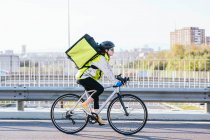 Side view of female courier with thermal bag riding bike on bridge while delivering food in city — Stock Photo