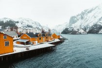 Желтые коттеджи и заснеженная набережная, расположенная рядом с рябью моря на фоне гор в холодный зимний день в прибрежной деревне на Лофских островах, Норвегия — стоковое фото