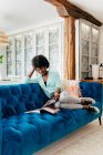 Молодая черная женщина в повседневной одежде с наушниками сидит на уютном синем диване дома и читает журнал, наслаждаясь свободным временем дома — стоковое фото