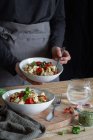 Cultivo pessoa anônima cozinhar saborosa massa assada com metades de tomate cereja e folhas de manjericão fresco em tigelas — Fotografia de Stock