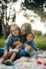 Feliz madre joven con lindas hijas con ropa similar disfrutando de un soleado día de verano juntas mientras están sentadas en una manta en un césped cubierto de hierba en el parque - foto de stock