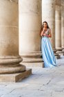 Полное тело очаровательной молодой модели в элегантном модном синем вечернем платье, стоящем рядом со старыми колоннами здания, смотрящим в камеру — стоковое фото