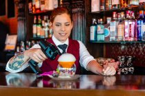 Skilled young female barkeeper using flavor bluster smoke gun while garnishing cocktail at bar counter — Stock Photo