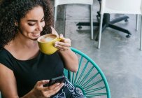 De cima de feliz jovem hispânico feminino navegando telefone celular enquanto desfruta de delicioso cappuccino na mesa de café de rua — Fotografia de Stock