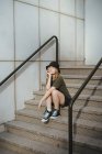 Junge Frau in lässiger Kleidung blickt in die Kamera, die tagsüber auf einer Treppe an der Betonwand eines modernen Gebäudes in der Stadtstraße sitzt — Stockfoto