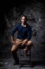 Delighted stylish ethnic male sitting on wooden stool in studio on dark background and looking at camera — Stock Photo