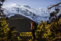 Vista laterale dello zaino viaggiatore ammirando magnifico paesaggio di boschi di conifere che crescono sullo sfondo delle montagne innevate dell'Himalaya sotto il cielo blu nella giornata di sole in Nepal — Foto stock