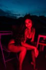 Молода жінка в нижній білизні дивиться на камеру, сидячи на стільці під червоним неоновим світлом вночі на терасі — стокове фото