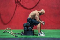 Vista lateral do homem musculoso exausto olhando para longe sentado em pesos e descansando durante o treino funcional no ginásio — Fotografia de Stock