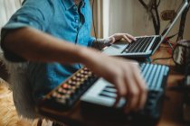 Side view of unrecognizable cropped young man working on synthesizer and laptop at table at home — Stock Photo