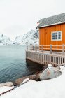 В холодный зимний день в прибрежной деревушке на Лофотенских островах, Норвегия, рядом с рябью моря на фоне гор стоит желтый кабан и заснеженная набережная — стоковое фото