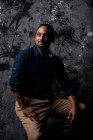 Serious stylish ethnic male sitting on wooden stool in studio on dark background and looking away — Stock Photo