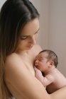 Vista laterale di tenera madre in topless con gli occhi chiusi in piedi con carino neonato nudo vicino al muro a casa — Foto stock
