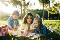 Glückliche junge Mutter mit kleinen Töchtern, die auf einer Decke liegen und spielen, während sie den Sommertag zusammen im sonnigen Park verbringen — Stockfoto