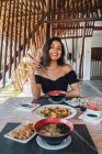 Alegre turista femenina con deliciosa pasta entre palitos de comida por encima de la mesa con salsa de soja y rodajas de jengibre en vinagre al aire libre - foto de stock
