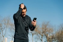 Бородатый спортсмен в спортивной одежде и наушниках просматривает мобильный телефон, стоя на лугу в городе — стоковое фото