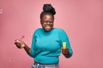 Heureuse femme afro-américaine avec les yeux fermés portant des vêtements bleus et soufflant des bulles de savon sur fond rose en studio — Photo de stock