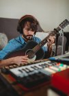 Young man in headphones playing on guitar near table with laptop and synthesizer at home looking at camera — Stock Photo