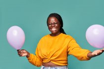 Eccitato afroamericano femminile in jeans e top giallo in piedi vicino caduta palloncini contro sfondo blu in studio — Foto stock