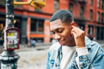 From below cheerful content African American guy in denim trendy jacket surfing modern mobile phone during city Side view of cheerful content African American guy in denim trendy jacket putting on headphones during city stroll — Stock Photo