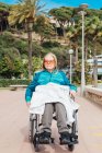 Неемоційна літня жінка в інвалідному візку катається по набережній біля моря і насолоджується літнім днем — стокове фото