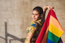 Серьезные молодые бисексуальные этнические женщины с разноцветным флагом, представляющим символы ЛГБТК и смотрящие в камеру в солнечный день — стоковое фото