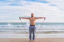 Shirtless brutal man looking at camera doing side lateral flies with resistant band training on seashore and looking forward — Stock Photo