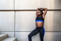 Pretty black female in sportswear keeping eyes closed and bending aside while doing warming up exercises near wall of modern building on city street — Stock Photo