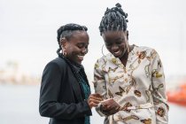 Trendige lächelnde Afroamerikanerinnen mit Frisur verbringen bei hellem Tag Zeit miteinander beim Handy-Surfen im Park — Stockfoto