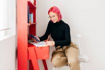 Вид збоку молодої творчої жінки-дизайнера з рожевим волоссям у повсякденному вбранні та окулярах за допомогою смартфона та ескізу малювання під час роботи за столом вдома — стокове фото