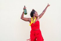 Mulher afro-americana encantada em colar de flores vestindo brilhante posição geral com garrafa de champanhe contra fundo branco — Fotografia de Stock