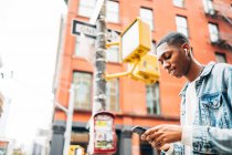 From below cheerful content African American guy in denim trendy jacket surfing modern mobile phone during city stroll — Stock Photo