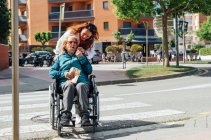 Mujer adulta empujando silla de ruedas con madre mayor y cruzando la calle en la ciudad durante el paseo en verano - foto de stock