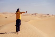 Vista lateral de una joven sonriente descalza vestida con ropa casual parada en una duna de arena contra el desierto y señalando hacia otro lado durante el viaje en Emiratos - foto de stock