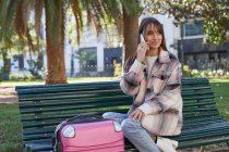 Позитивная молодая женщина-путешественница в стильном пальто сидит на скамейке возле чемодана и разговаривает по мобильному телефону в городском парке весной — стоковое фото