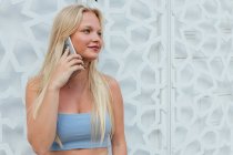 Mulher sorridente calma com cabelo loiro e em roupa de verão em pé na cidade e falando no telefone celular enquanto olha para longe — Fotografia de Stock