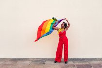 Femme afro-américaine joyeuse dans des vêtements élégants avec bouteille de boisson alcoolisée et drapeau coloré regardant vers le haut sur fond clair — Photo de stock