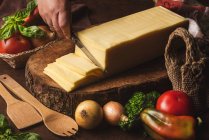 Bloco de queijo em suporte de madeira perto de cebolas cruas e saco de tecelagem contra espátulas orgânicas e folhas de manjericão — Fotografia de Stock