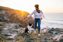 Young African American female owner running with Border Collie dog while spending time together on beach near waving sea at sunset — Stock Photo