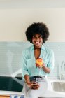 Moderne schöne Afroamerikanerin mit Smartphone in der Hand sitzt auf dem Küchentisch und schaut in die Kamera, lächelt zu Hause und isst Apfel — Stockfoto