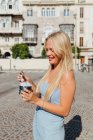 Beautiful blonde young female eating cold tasty ice cream while standing in city street on sunny day in summer — Stock Photo