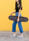 Обрезанный неузнаваемая молодая фигуристка со скейтбордом стоя глядя в сторону на дорожке с красочной желтой стеной на заднем плане в дневное время — стоковое фото
