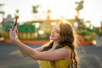 Content female taking self portrait on mobile phone while having fun in amusement park in evening in summer — Stock Photo