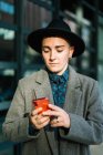 Androgynous pessoa em chapéu navegando no celular olhando para a tela de pé na rua à luz do dia — Fotografia de Stock