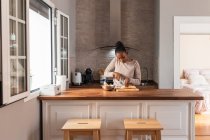 Schwarze Frau in Kleidung mit gestreiftem Ornament schneidet Kochbanane mit Messer auf Schneidebrett am Tisch im Haus — Stockfoto