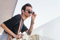 Low angle side view of gay male in trendy sunglasses leaning on glass railing on balcony and looking away — Stock Photo